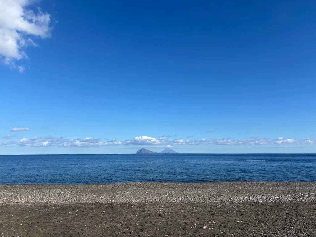 A relaxing view from Canneto beach in Lipari, showing the dark volcanic pebbles and the smoking Stromboli volcano on the horizon during the Riding into the Lava tour