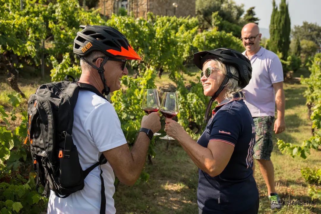 Two mountain bikers toasting with glasses of red wine in a lush vineyard during a Trails Unlimited guided tour in the Langhe region.