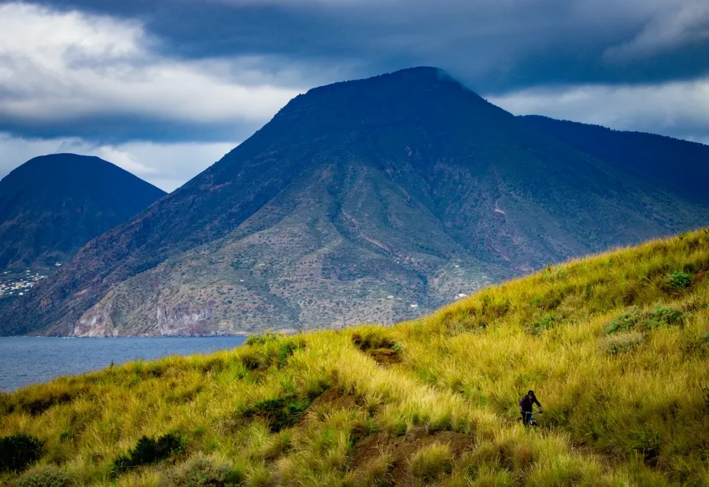 Mountain biker riding through golden fields on a volcanic trail in Lipari, Sicily, with a stunning view of the Aeolian islands during a Trails Unlimited tour.
