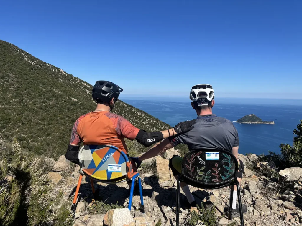 Two mountain bikers sitting on chairs and relaxing on a rocky viewpoint overlooking the Mediterranean Sea and the Bergeggi island, while a group of kids and their guide ride mountain bikes on a trail below during a Trails Unlimited Family Camp in Finale Ligure.