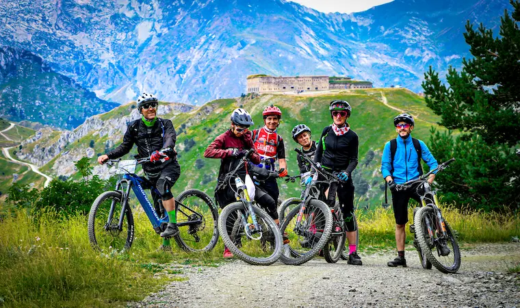 A group of mountain bikers smiling and standing with their bikes on a dirt path during a guided Trails Unlimited tour, with a historic stone fort and majestic green and blue mountains in the background.