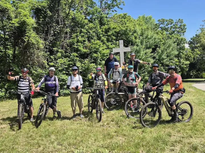 A group of mountain bikers posing with their e-bikes during an emtb langhe tour, standing on a grassy trail in front of a stone cross and lush green trees on a sunny day.