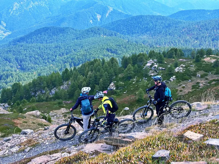 Three mountain bikers on a rocky trail along the Via del Sale, overlooking forested valleys in the Italian Alps.